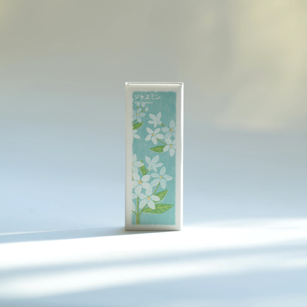 Kousaido Ikadenshin Japanese Incense Jasmine scent with a light blue front panel featuring white flowers and green leaves, standing upright on a white surface with soft sunlight and shadows.