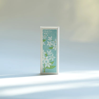 Kousaido Ikadenshin Japanese Incense Jasmine scent with a light blue front panel featuring white flowers and green leaves, standing upright on a white surface with soft sunlight and shadows.