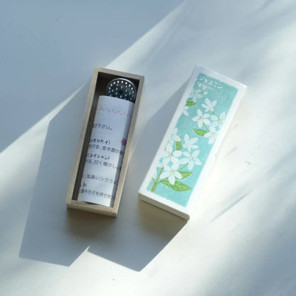 A small cylindrical Kousaido Ikadenshin Japanese Incense tube with Japanese text sits inside an open light wood box next to its rectangular lid, which has a mint green floral print. The scene is photographed on a white surface with strong diagonal shadows.