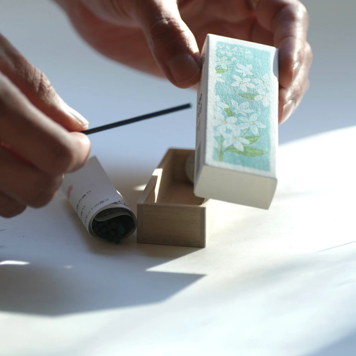 A close-up of hands holding a Kousaido Ikadenshin Japanese Incense with white floral artwork on a light blue background, while another hand holds a thin black rod over an open wooden box containing a rolled white item.