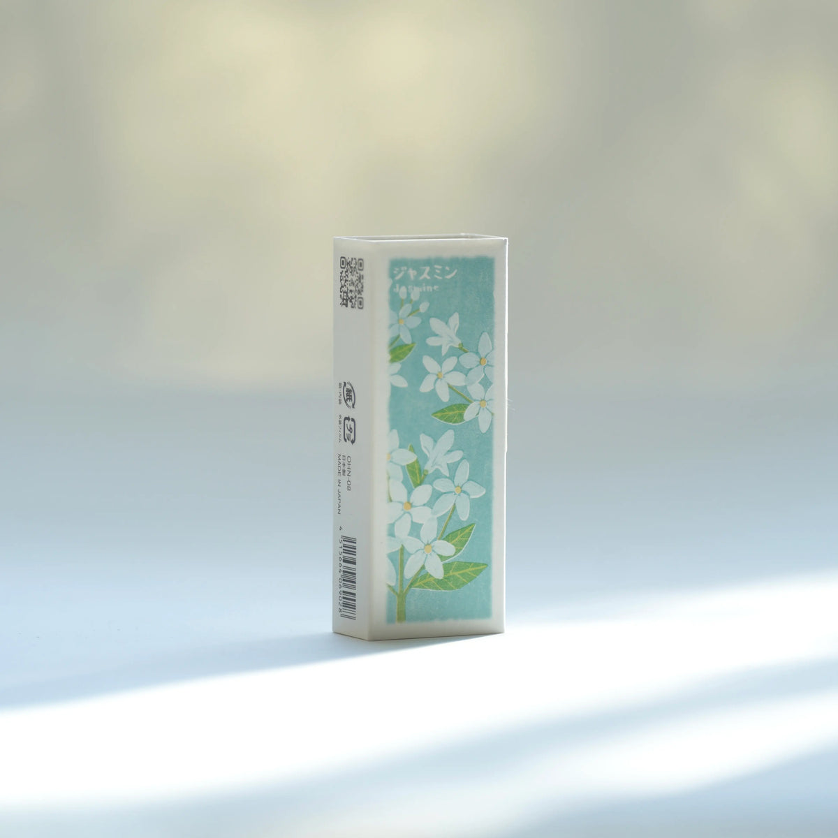 A Kousaido Ikadenshin Japanese Jasmine Incense stands upright on a light surface against a soft, bright background. The box is white with a light blue front panel decorated with white flowers and green leaves.
