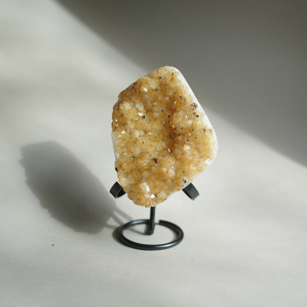 A raw crystal geode specimen with dense golden-yellow and white quartz druses, mounted on a small black metal display stand against a neutral light gray background with a soft shadow.
