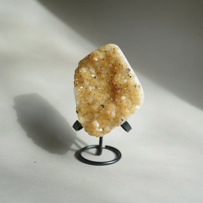 A raw crystal geode specimen with dense golden-yellow and white quartz druses, mounted on a small black metal display stand against a neutral light gray background with a soft shadow.
