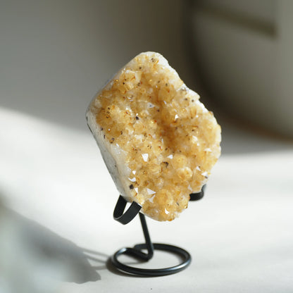 A natural crystal cluster geode specimen with golden-yellow and cream quartz points, displayed on a small black metal stand against a softly blurred light background.