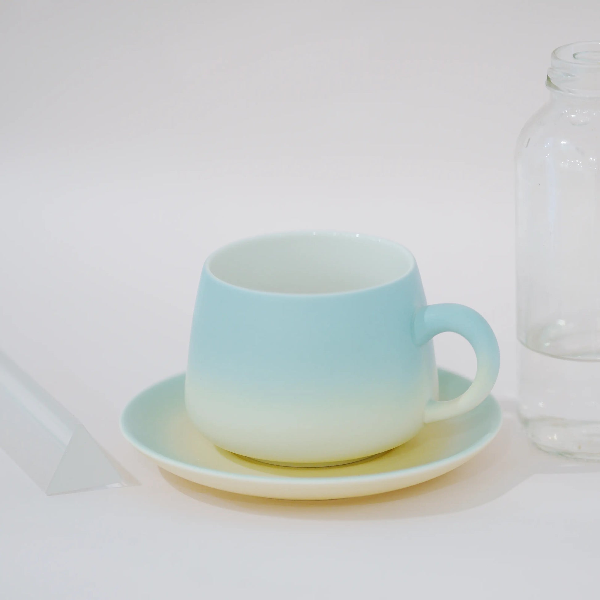 A ceramic cup and saucer set with a light blue to pale yellow gradient, shown on a white surface next to a clear glass bottle.