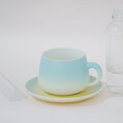 A ceramic cup and saucer set with a light blue to pale yellow gradient, shown on a white surface next to a clear glass bottle.