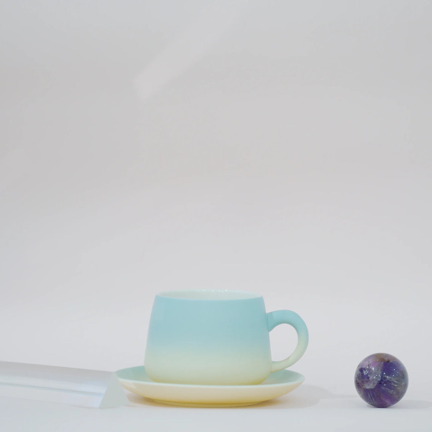 A ceramic teacup and saucer set with a smooth vertical gradient transitioning from light aqua blue at the top to soft cream at the bottom, styled on a white surface next to a small purple galaxy bead.