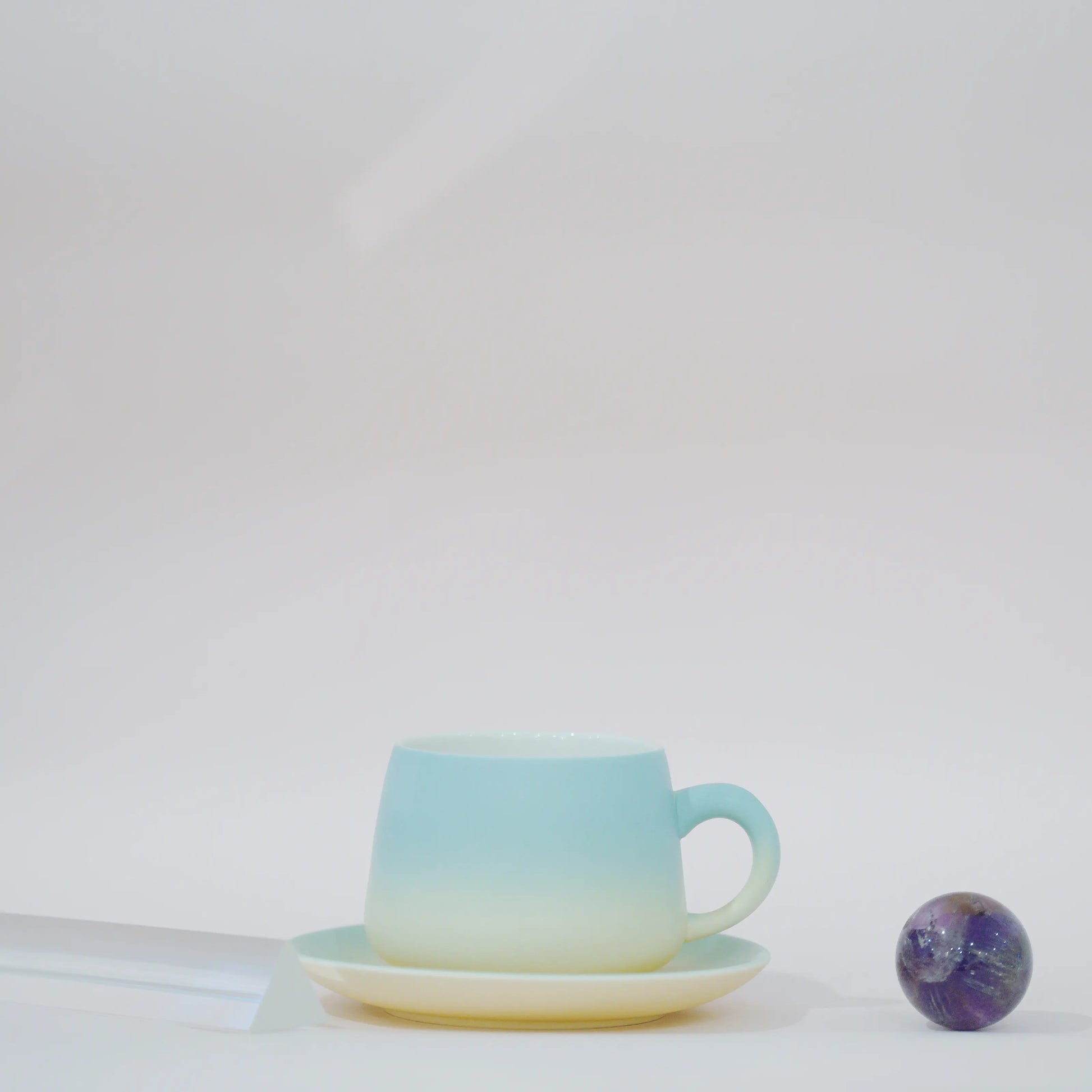 A ceramic teacup and saucer set with a smooth vertical gradient transitioning from light aqua blue at the top to soft cream at the bottom, styled on a white surface next to a small purple galaxy bead.