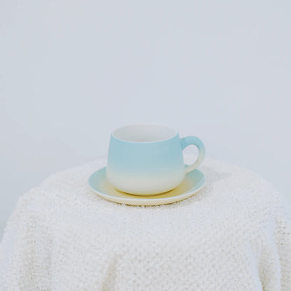 A pastel gradient ceramic cup and saucer set, featuring a soft blue-to-white transition with a subtle yellow accent on the saucer, displayed on a textured white knit blanket against a light gray background.