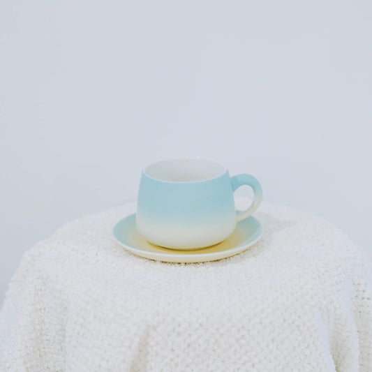 A pastel gradient ceramic cup and saucer set, featuring a soft blue-to-white transition with a subtle yellow accent on the saucer, displayed on a textured white knit blanket against a light gray background.