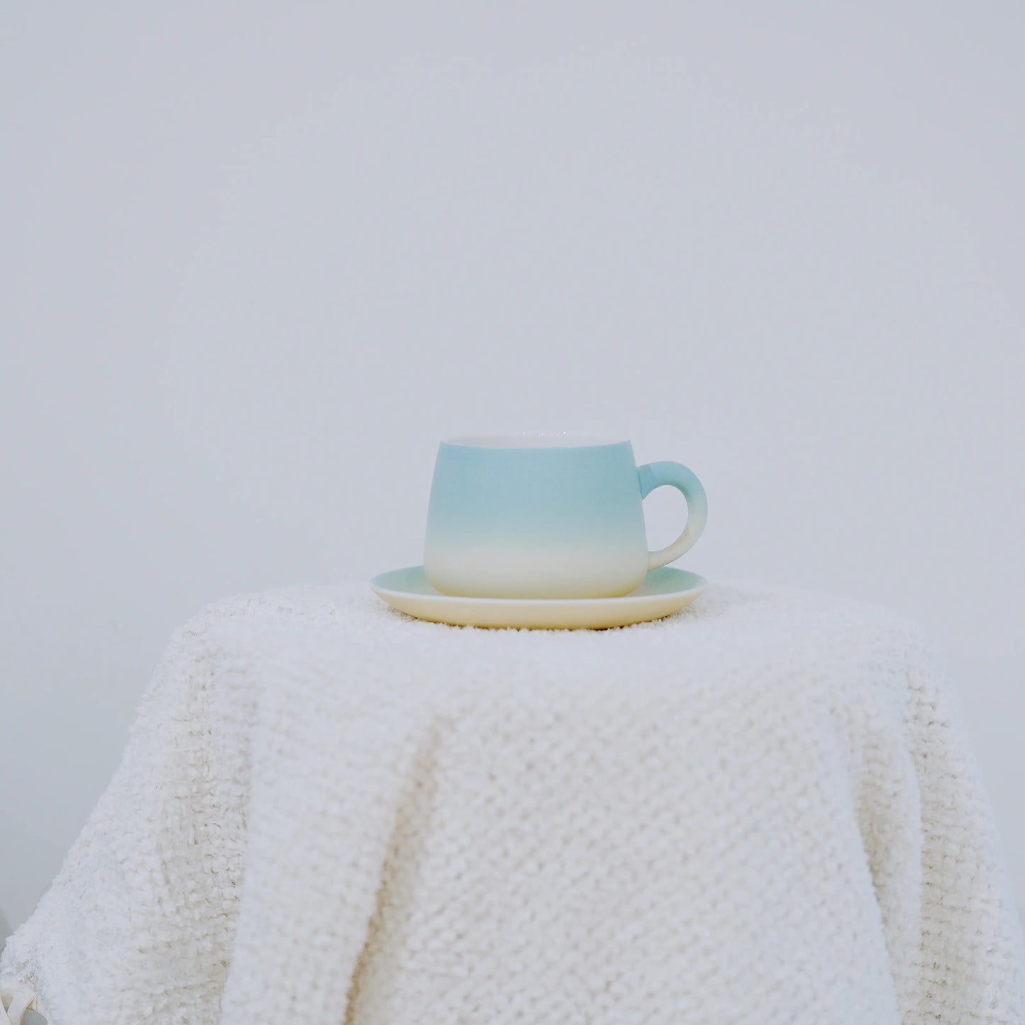 A ceramic cup and saucer set with a light blue to cream gradient, displayed on a textured white knit blanket against a plain light gray background.