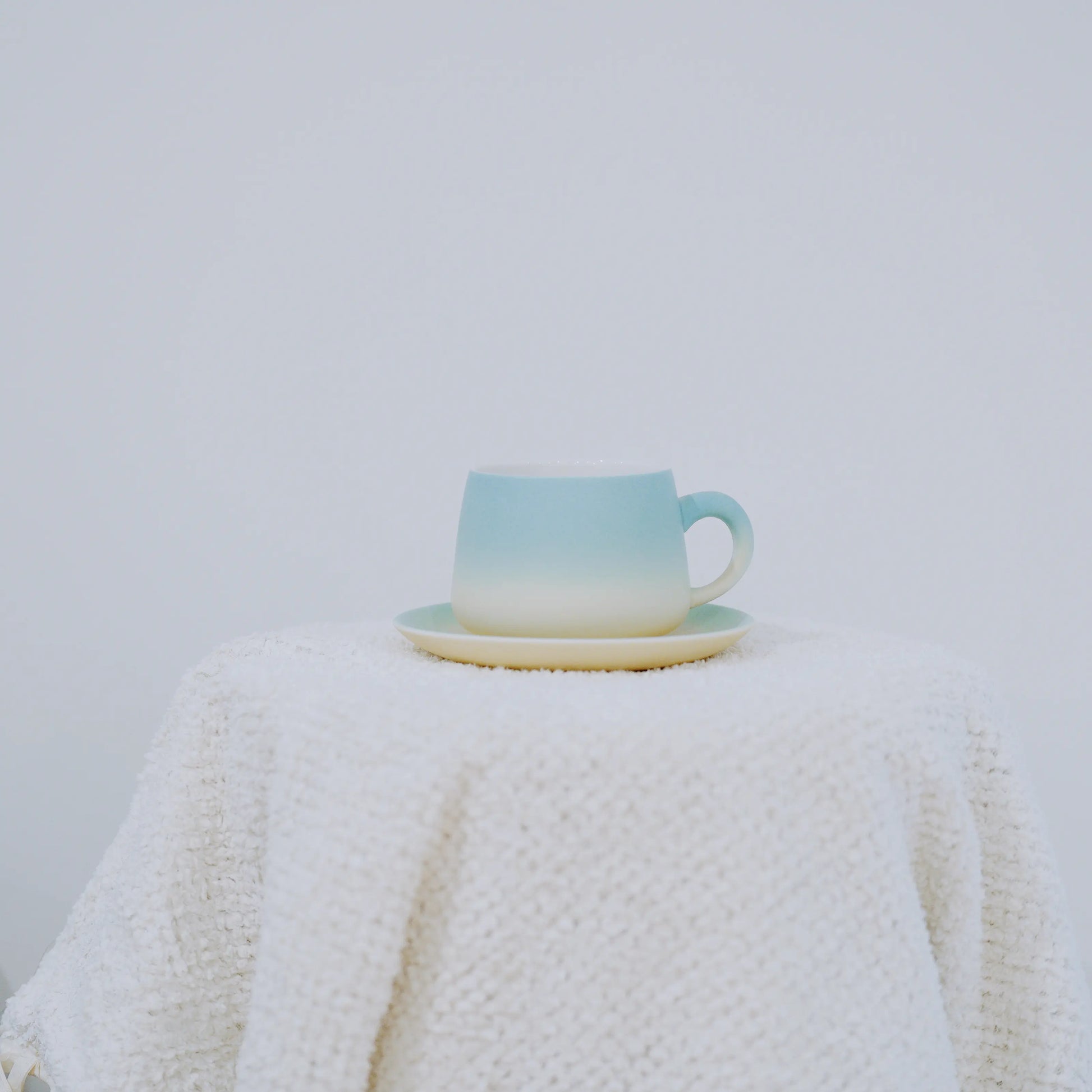 A ceramic cup and saucer set with a light blue to cream gradient, displayed on a textured white knit blanket against a plain light gray background.
