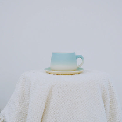 A ceramic cup and saucer set with a light blue to cream gradient, displayed on a textured white knit blanket against a plain light gray background.