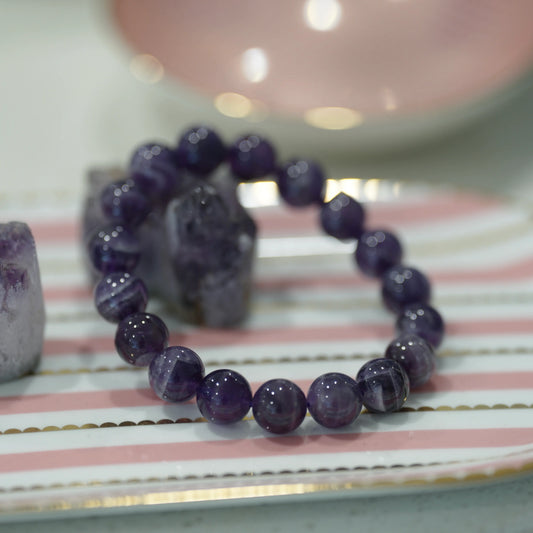 A Natural Uruguayan Dream Amethyst Crystal Bracelet 11mm displayed on a pink and white striped surface with crystals in the background.