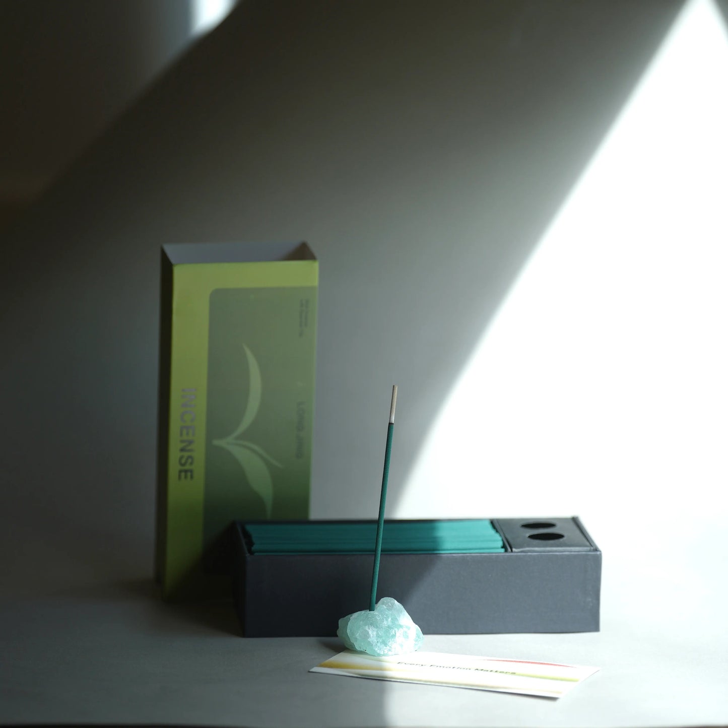 A minimalist incense set on a gray surface with dramatic diagonal light and shadow. The set includes a tall lime green and gray incense box, a dark charcoal incense holder with teal incense sticks, a single lit incense stick resting on a pale blue stone, and a small card.