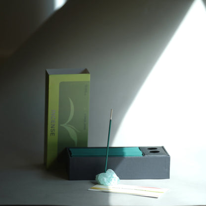 A minimalist incense set on a gray surface with dramatic diagonal light and shadow. The set includes a tall lime green and gray incense box, a dark charcoal incense holder with teal incense sticks, a single lit incense stick resting on a pale blue stone, and a small card.