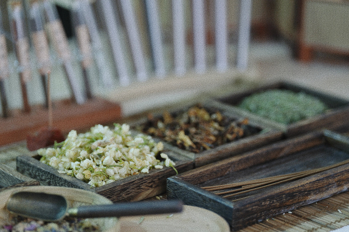 A close-up, shallow depth-of-field view of a wooden incense burner tray filled with dried herbs and incense sticks, with a scoop in the foreground and a blurred background.