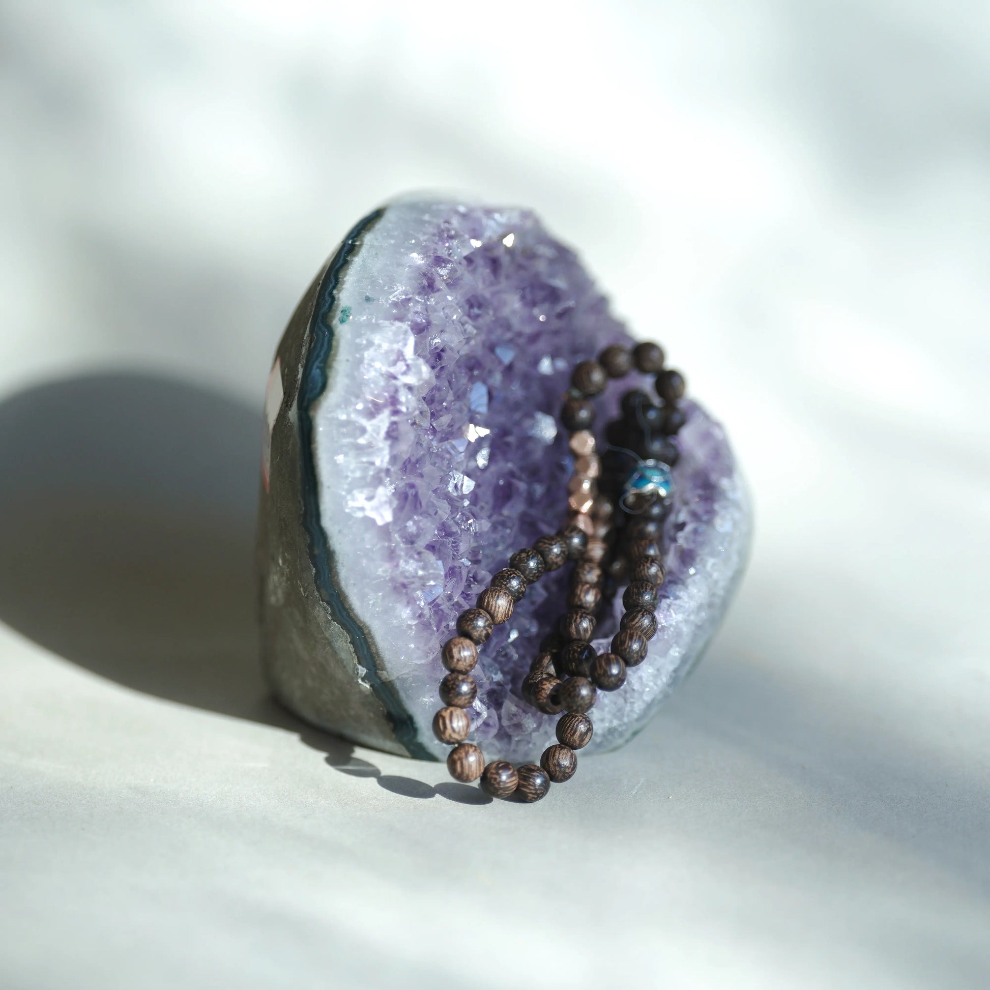A beaded necklace with dark brown wooden beads and a small blue bead accent, draped over a purple amethyst geode on a light beige surface.