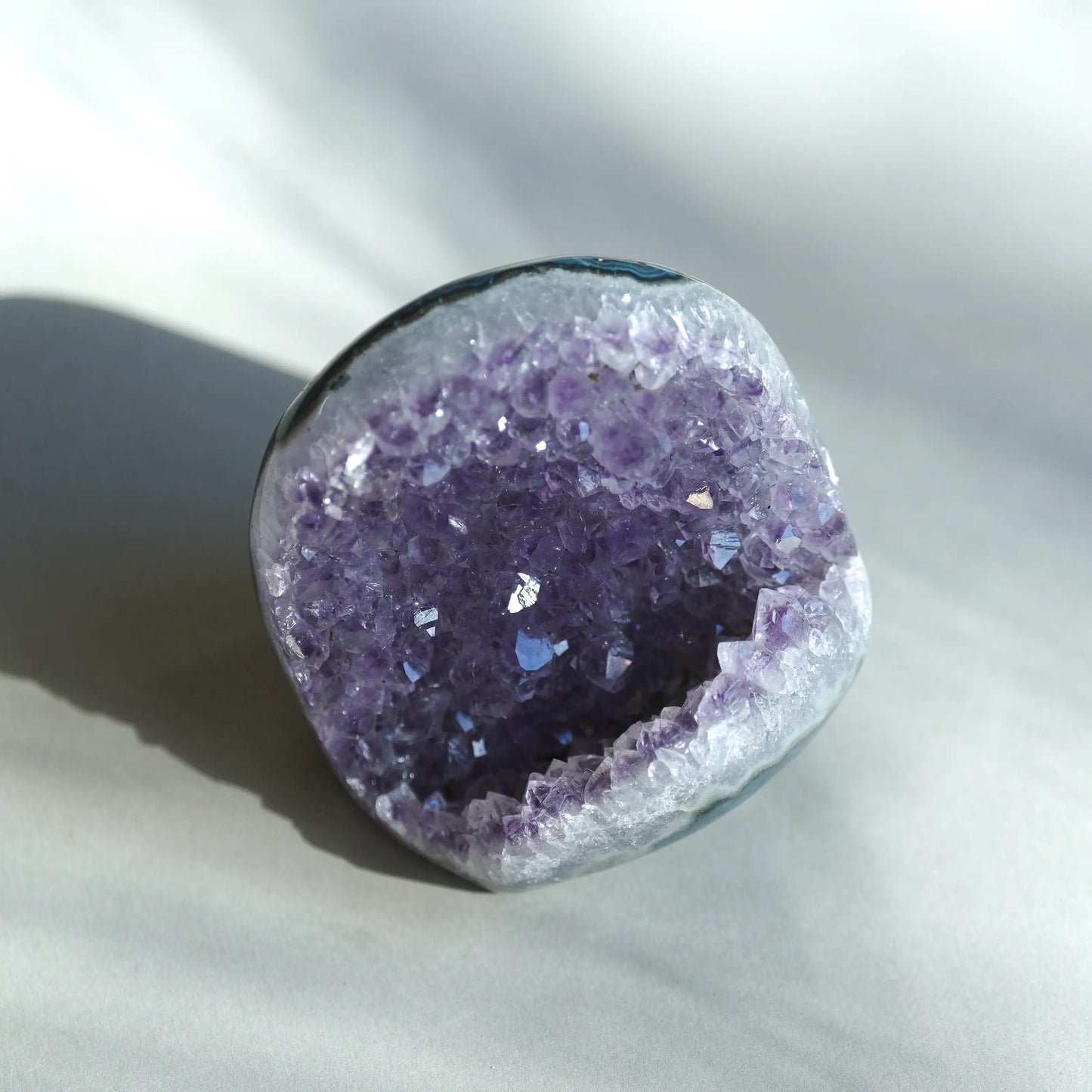 A raw amethyst geode specimen with a deep purple crystal interior and a smooth, polished gray outer rind, photographed on a light gray fabric background with soft shadows.