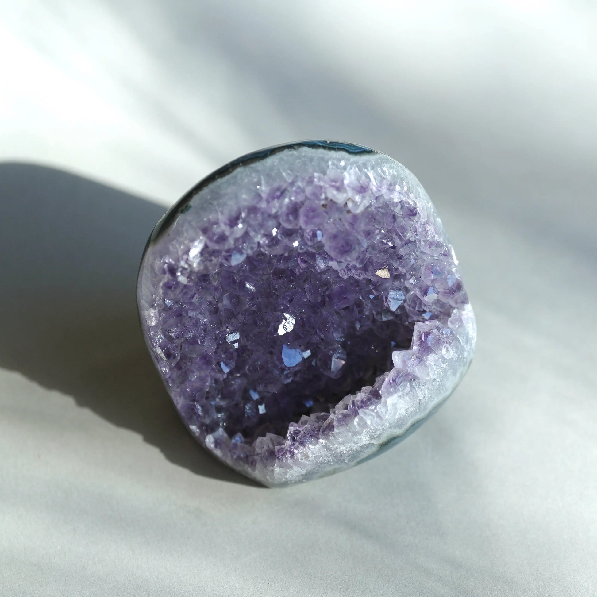 A raw amethyst geode specimen with a deep purple crystal interior and a smooth, polished gray outer rind, photographed on a light gray fabric background with soft shadows.