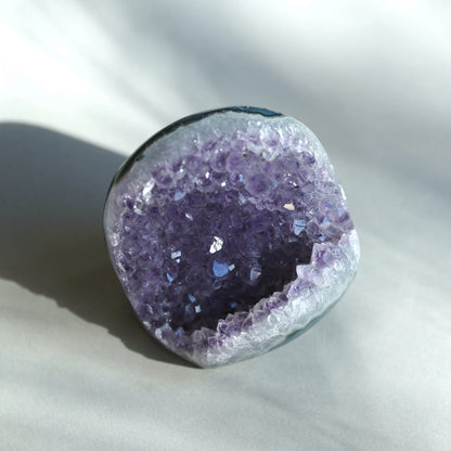 A raw amethyst geode specimen with a deep purple crystal interior and a smooth, polished gray outer rind, photographed on a light gray fabric background with soft shadows.