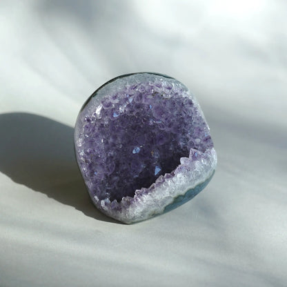 A polished amethyst geode specimen with a deep purple crystal interior and a white crystalline outer rim, photographed on a soft white fabric surface with a cast shadow.