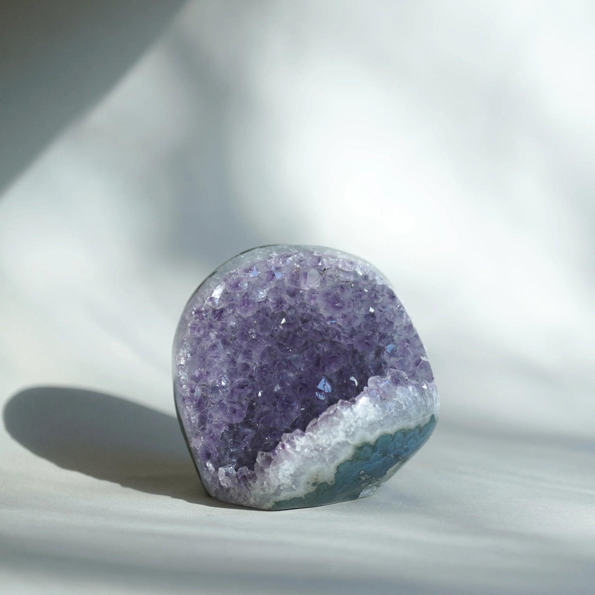 A polished purple amethyst geode crystal money bag with a white and blue crystalline base, photographed on a soft white fabric background with natural shadows.