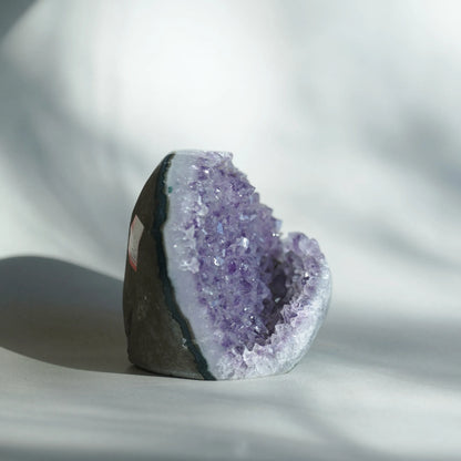 A raw amethyst geode specimen with a dark gray outer matrix and a prominent purple crystal cavity, photographed on a white draped fabric background with soft shadows.