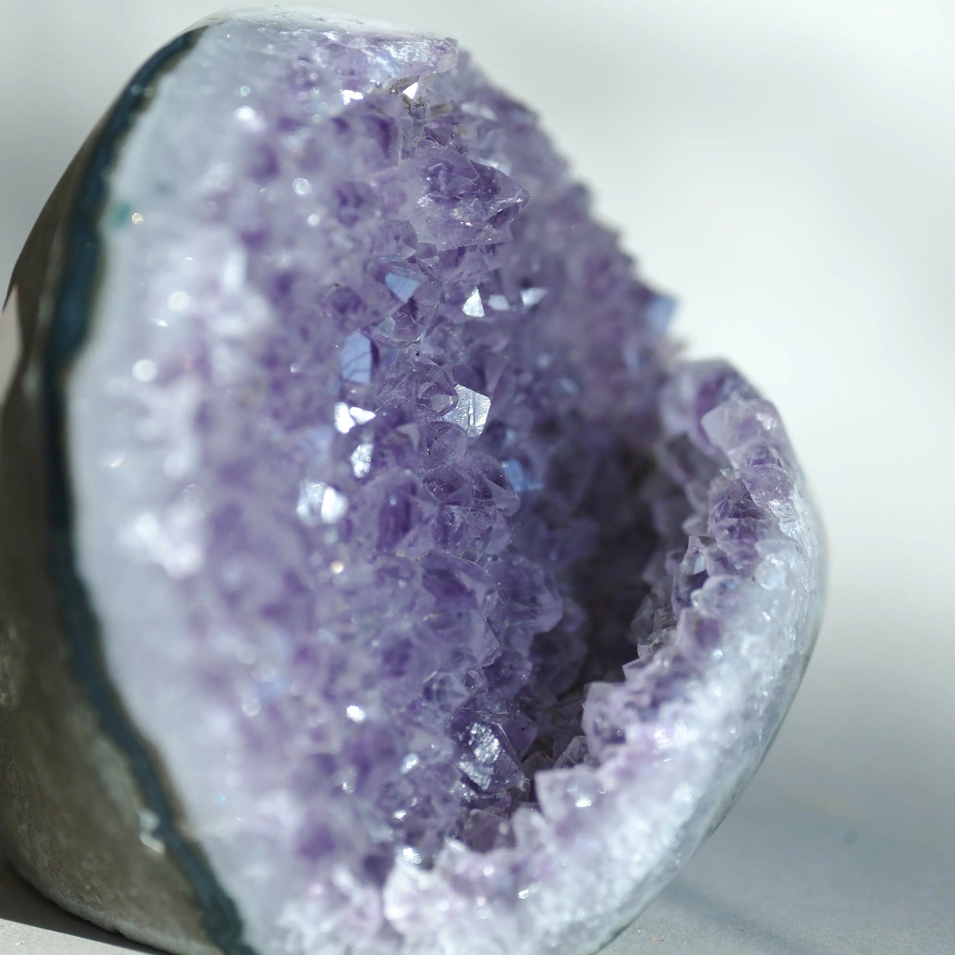 A close-up of a geode or crystal dome with a purple amethyst interior and a gray outer rind, photographed against a soft light gray background.