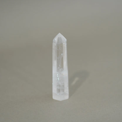 A natural clear quartz crystal point tower standing upright on a neutral gray surface with a soft shadow cast to the right.