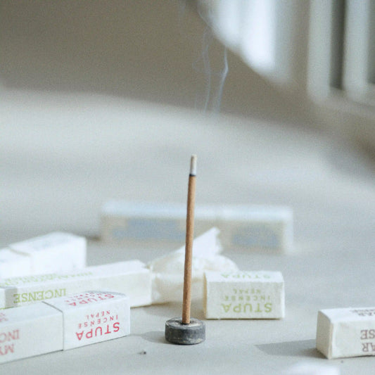A single incense stick stands upright in a small dark holder on a light surface, with several wrapped STUPA incense sticks scattered around it in a softly blurred indoor setting.