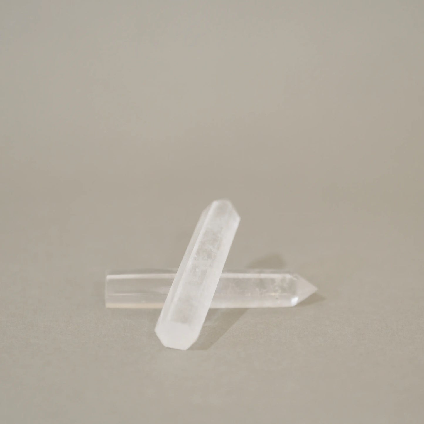 Two clear quartz crystal points crossed over each other on a neutral beige background.