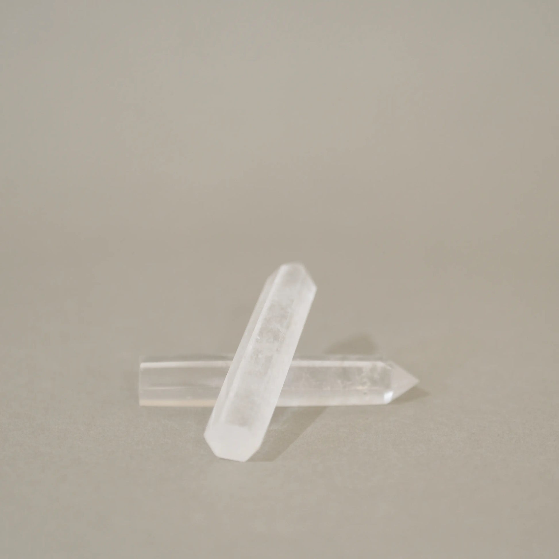 Two clear quartz crystal points crossed over each other on a neutral beige background.