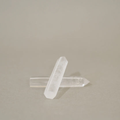 Two clear quartz crystal points crossed over each other on a neutral beige background.