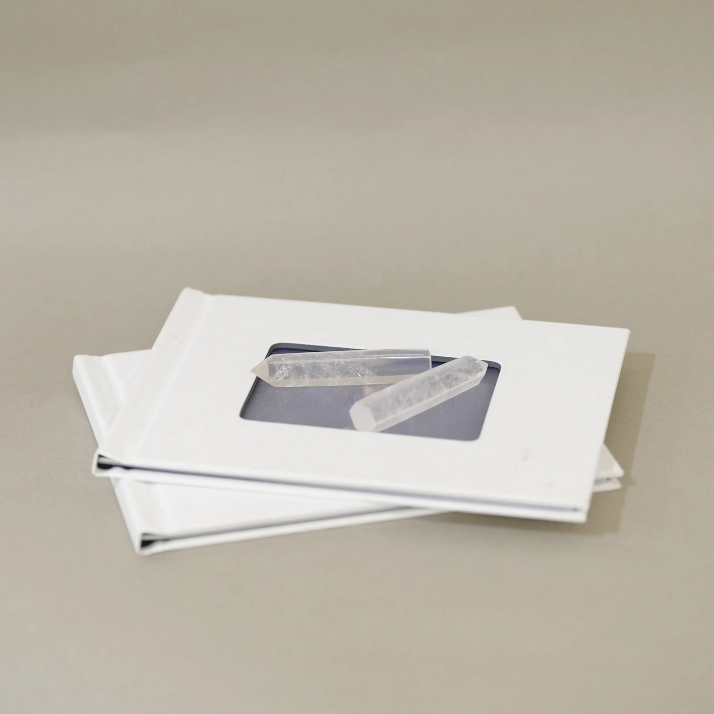 Two white square frames stacked on a neutral beige background, each with a small clear rectangular window showing a gray rectangular item inside.