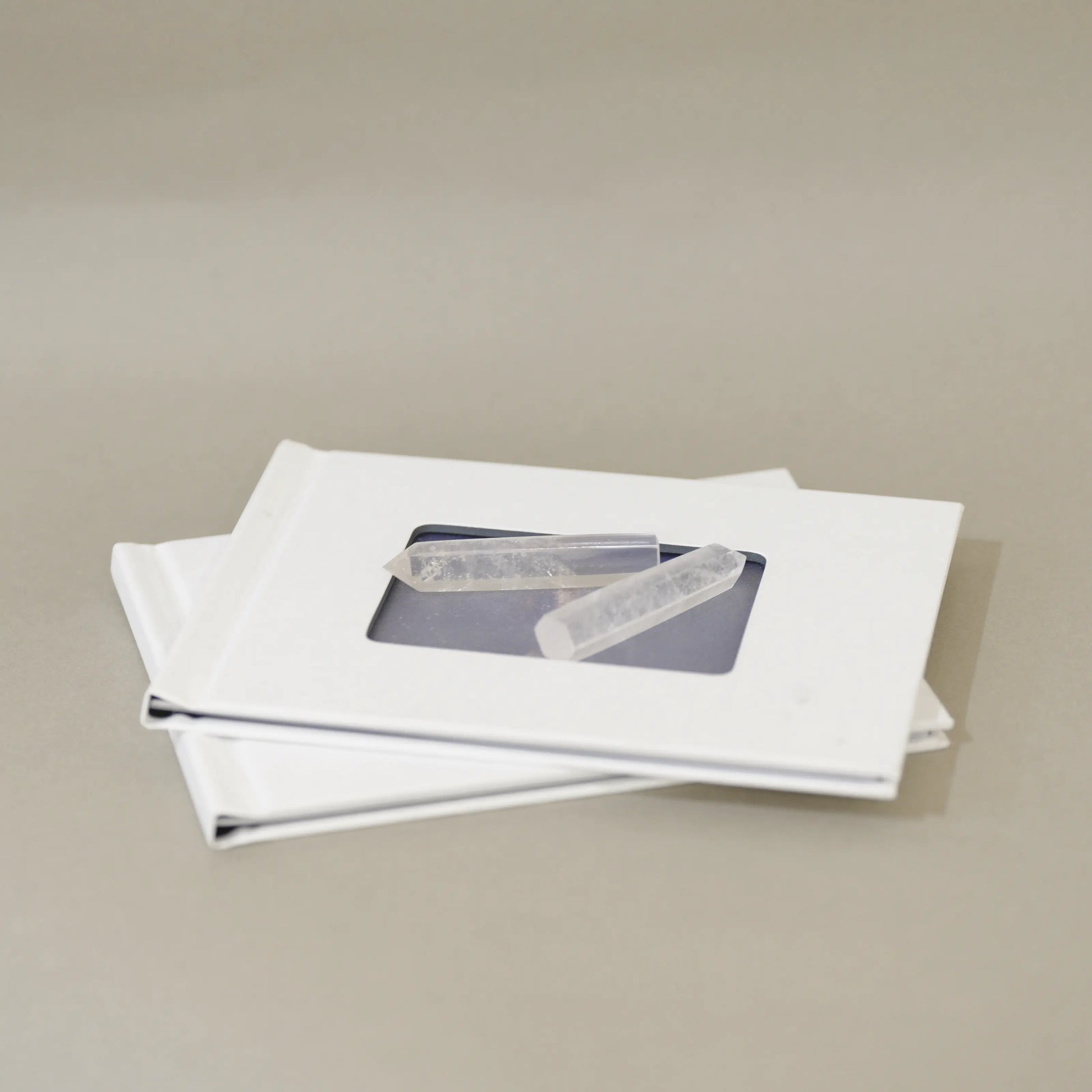 Two white square frames stacked on a neutral beige background, each with a small clear rectangular window showing a gray rectangular item inside.