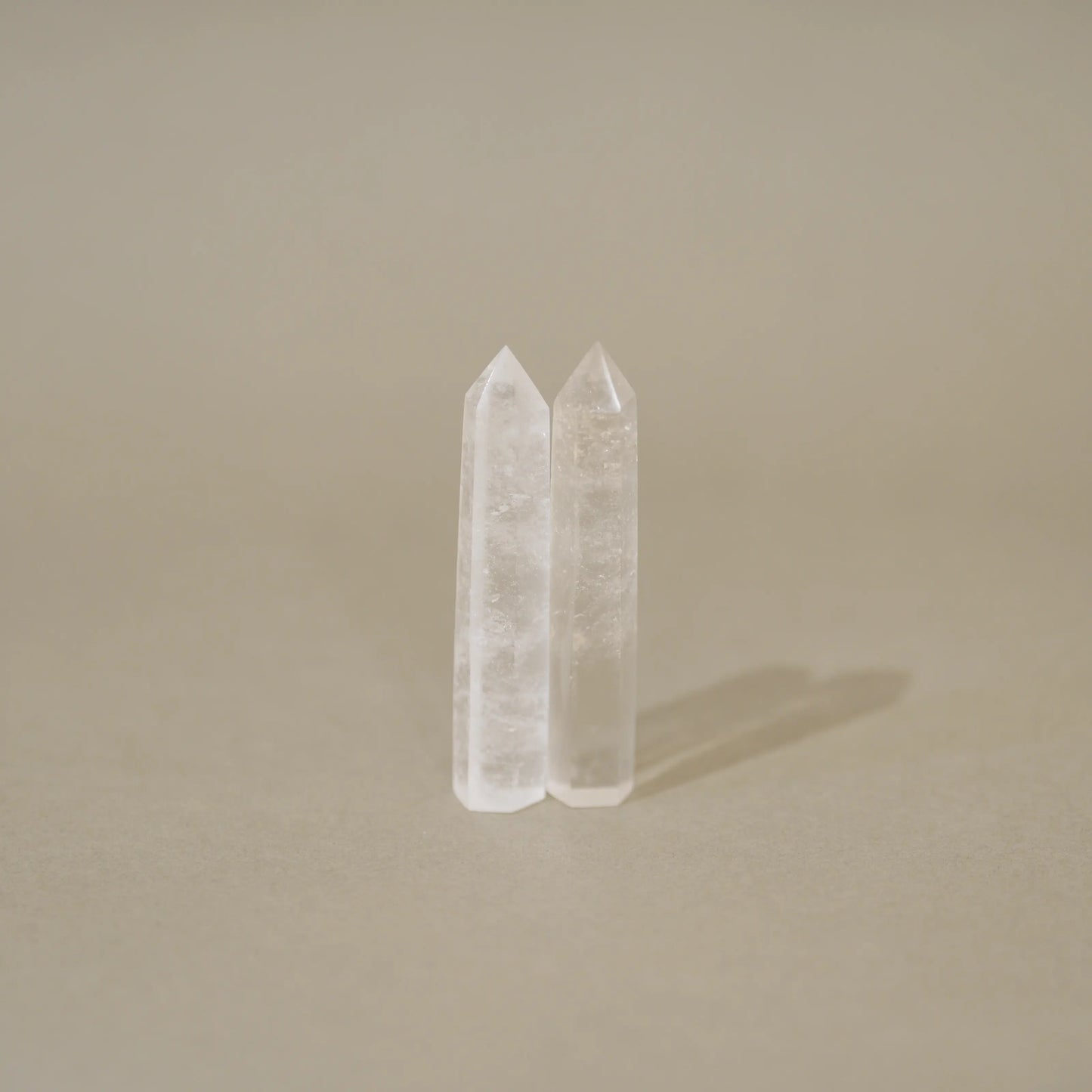 Two clear quartz crystal points standing upright on a neutral beige surface against a matching beige background.