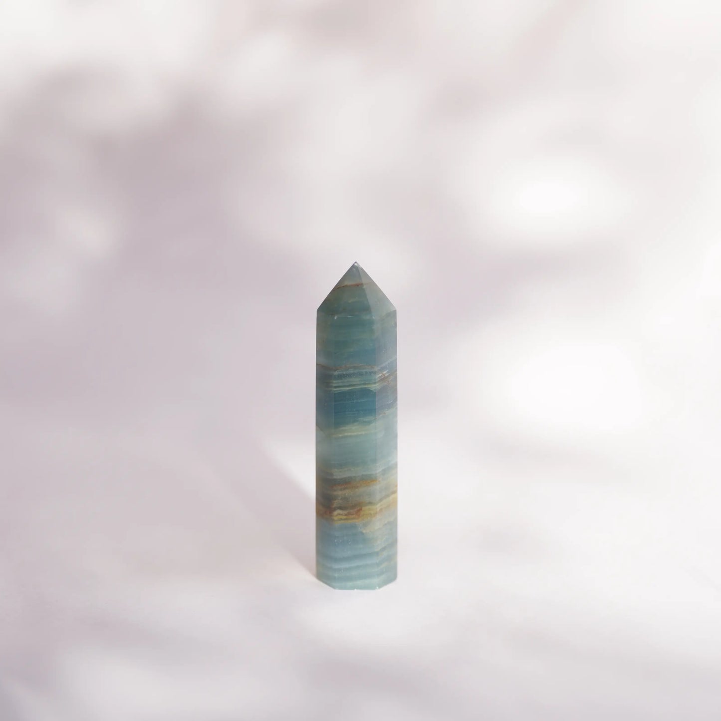A polished blue onyx crystal point tower with horizontal banding of light and dark layers, standing upright on a soft, light-colored surface with dappled shadows.