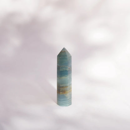 A polished blue onyx crystal point tower with horizontal banding of light and dark layers, standing upright on a soft, light-colored surface with dappled shadows.