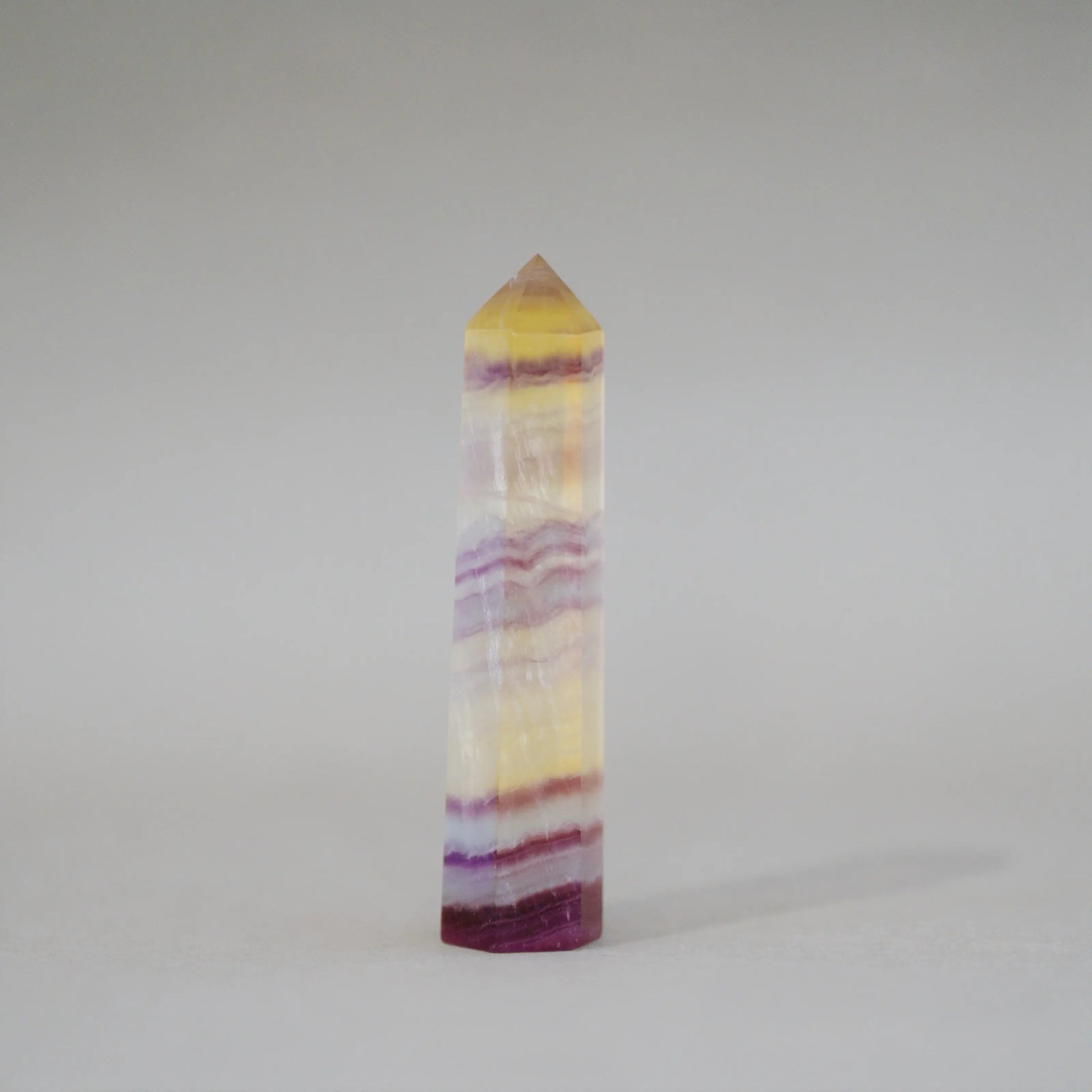 A natural Candy Fluorite Tower with horizontal bands of yellow, purple, and white, featuring a pointed pyramid top, photographed on a neutral light gray background.