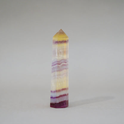 A natural Candy Fluorite Tower with horizontal bands of yellow, purple, and white, featuring a pointed pyramid top, photographed on a neutral light gray background.