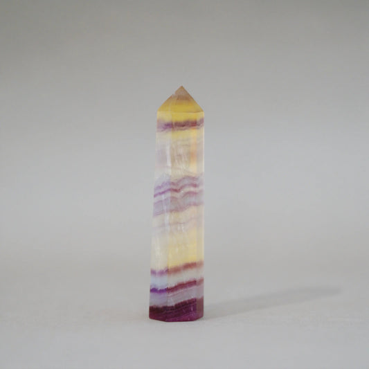 A natural Candy Fluorite Tower with horizontal bands of yellow, purple, and white, featuring a pointed pyramid top, photographed on a neutral light gray background.