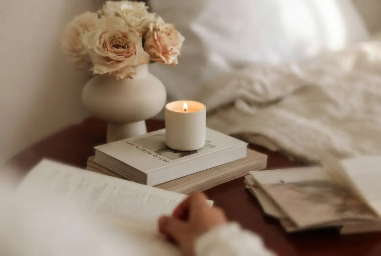 Cozy bedside scene with a lit candle on books, a white vase of pale roses, and a person reading in soft natural light.