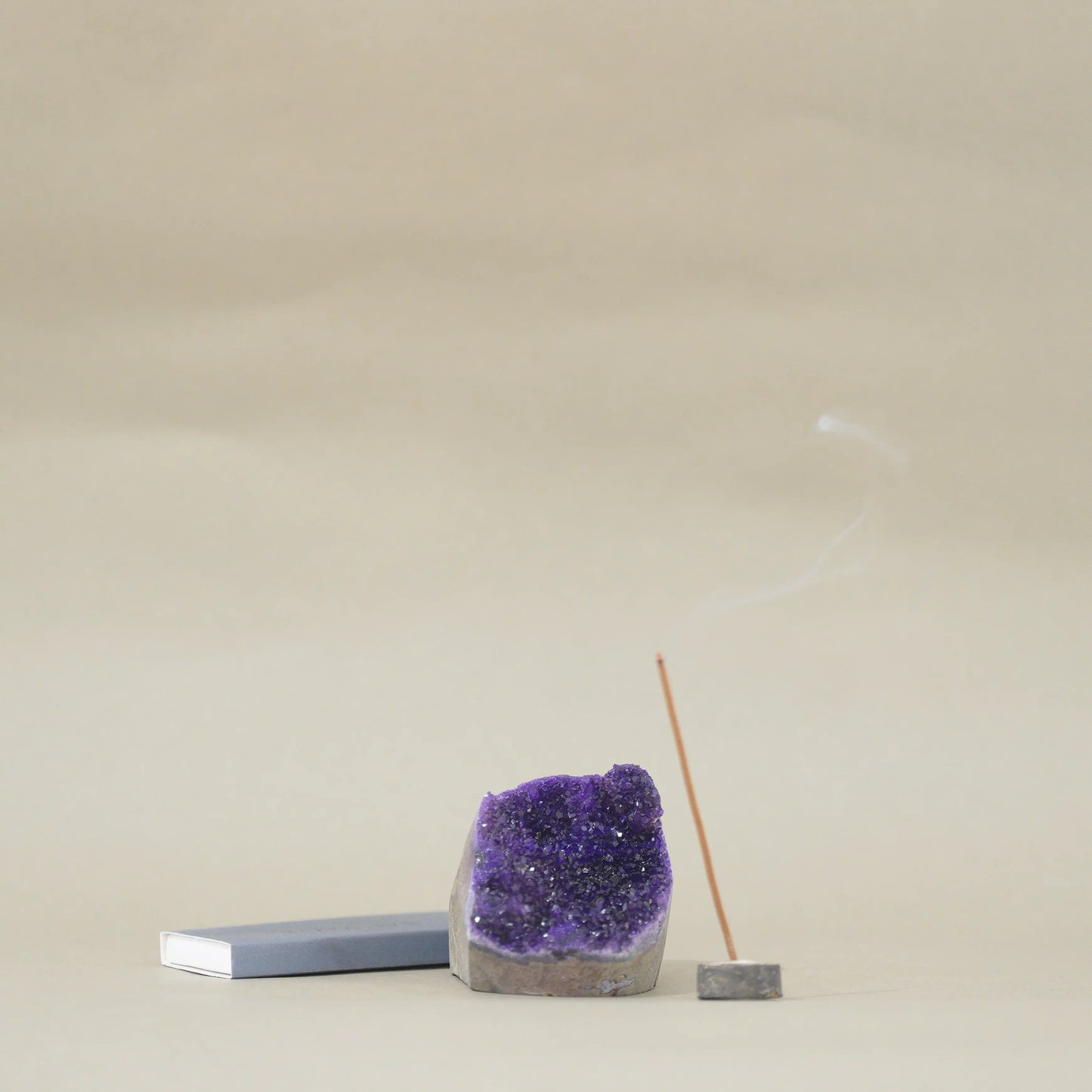 A purple amethyst geode crystal holder with a smoking incense stick on a small wooden base, next to a gray rectangular block, set against a neutral beige background.