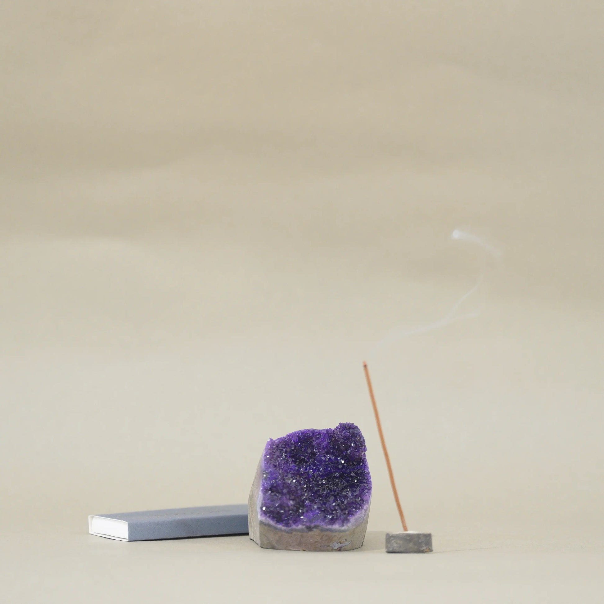 A purple amethyst geode crystal holder with a smoking incense stick on a small wooden base, next to a gray rectangular block, set against a neutral beige background.