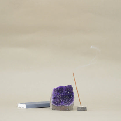 A purple amethyst geode crystal holder with a smoking incense stick on a small wooden base, next to a gray rectangular block, set against a neutral beige background.
