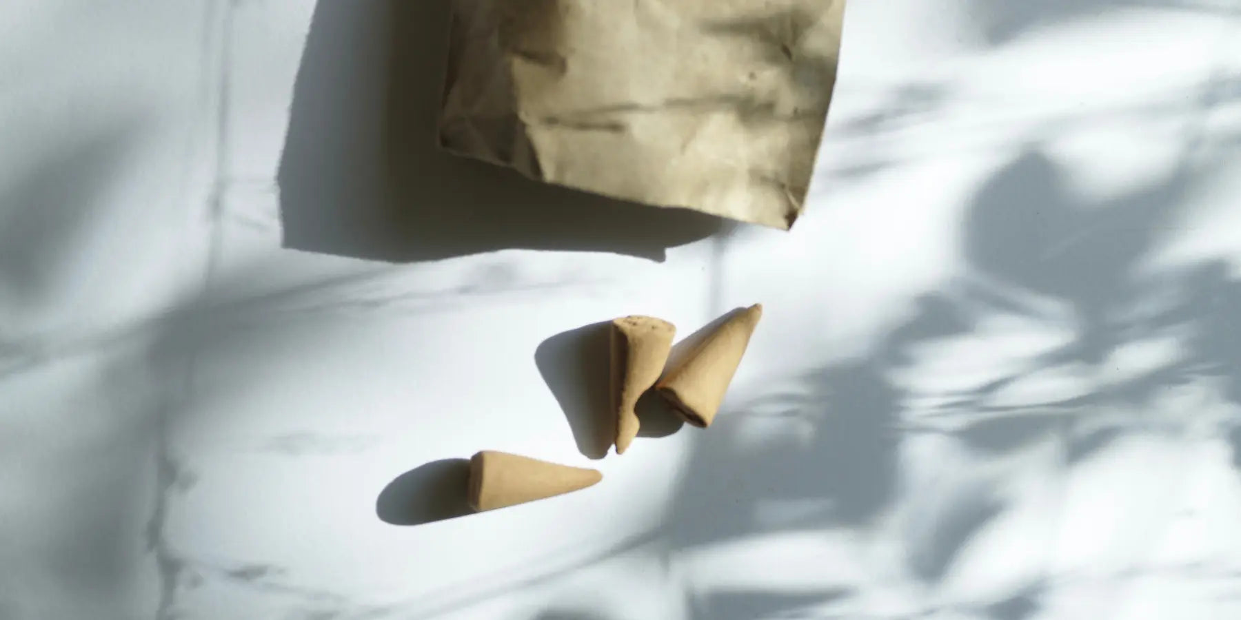 Three brown incense cones on a white surface with shadow patterns, near a brown paper bag.