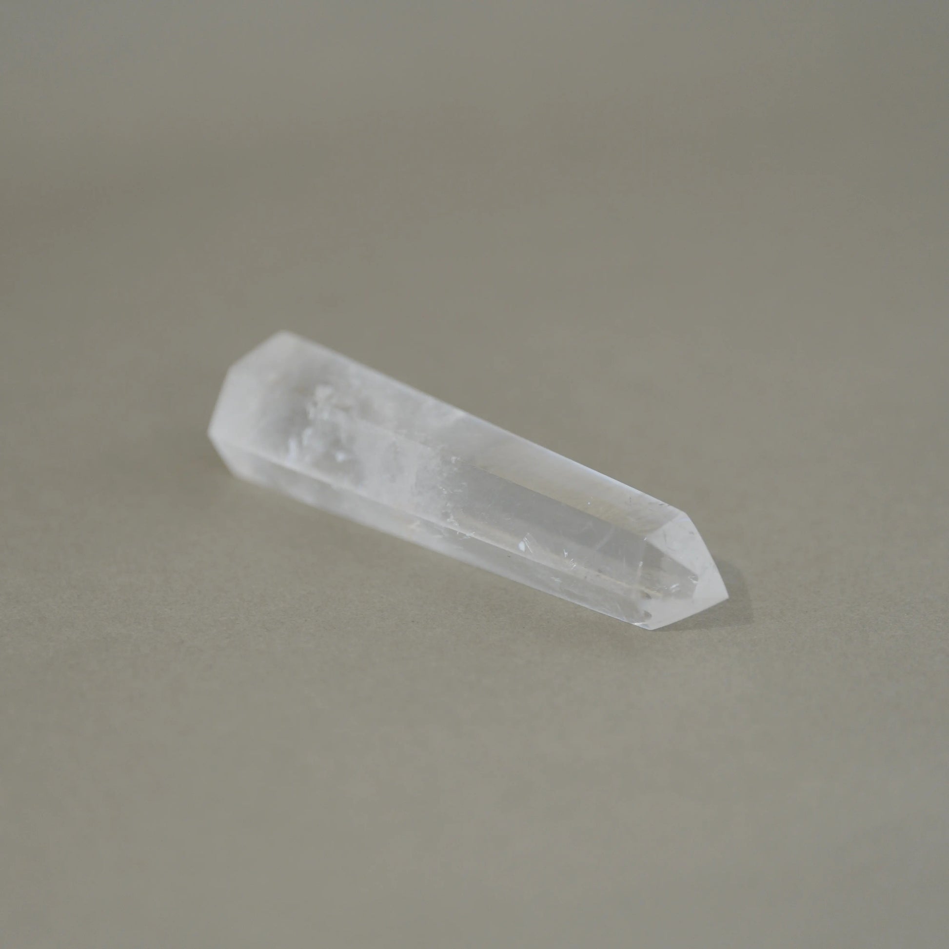 A natural clear quartz crystal point with a polished tip, resting on a smooth, solid taupe-gray surface.