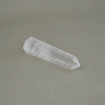 A natural clear quartz crystal point with a polished tip, resting on a smooth, solid taupe-gray surface.