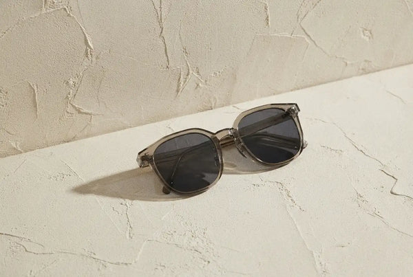 Transparent dark-framed sunglasses with black lenses resting on a textured beige surface.